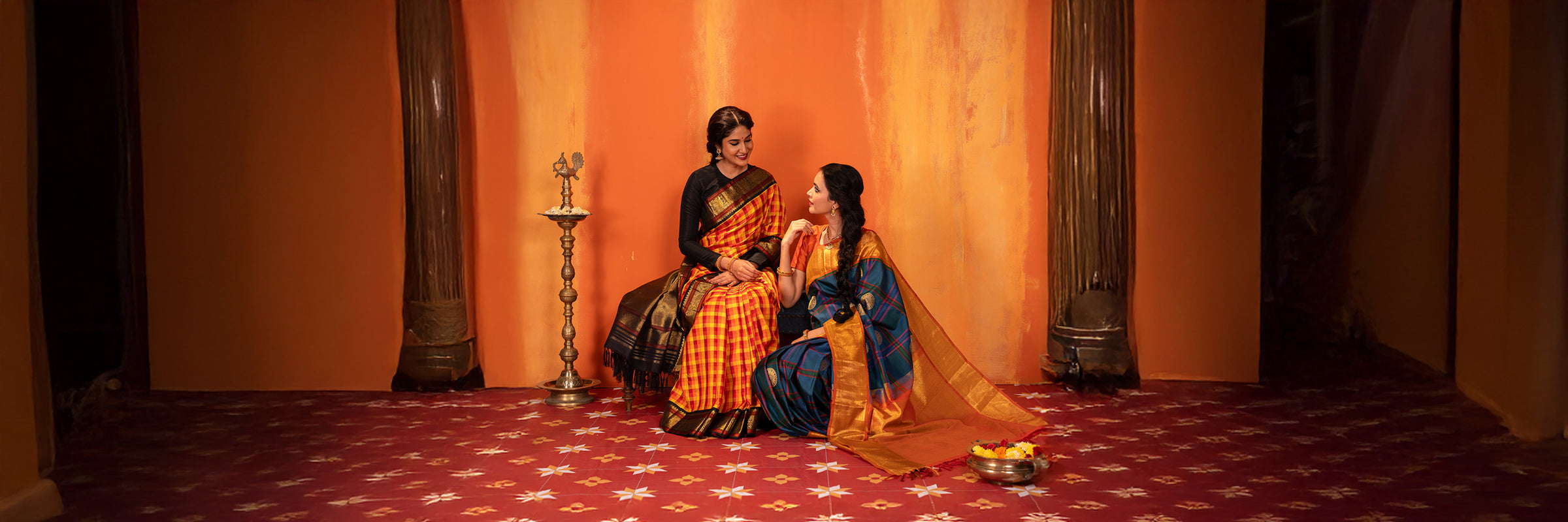 Two women in Vintage traditional kanjivaram Silks sitting on a patterned floor with an orange wall in the background.