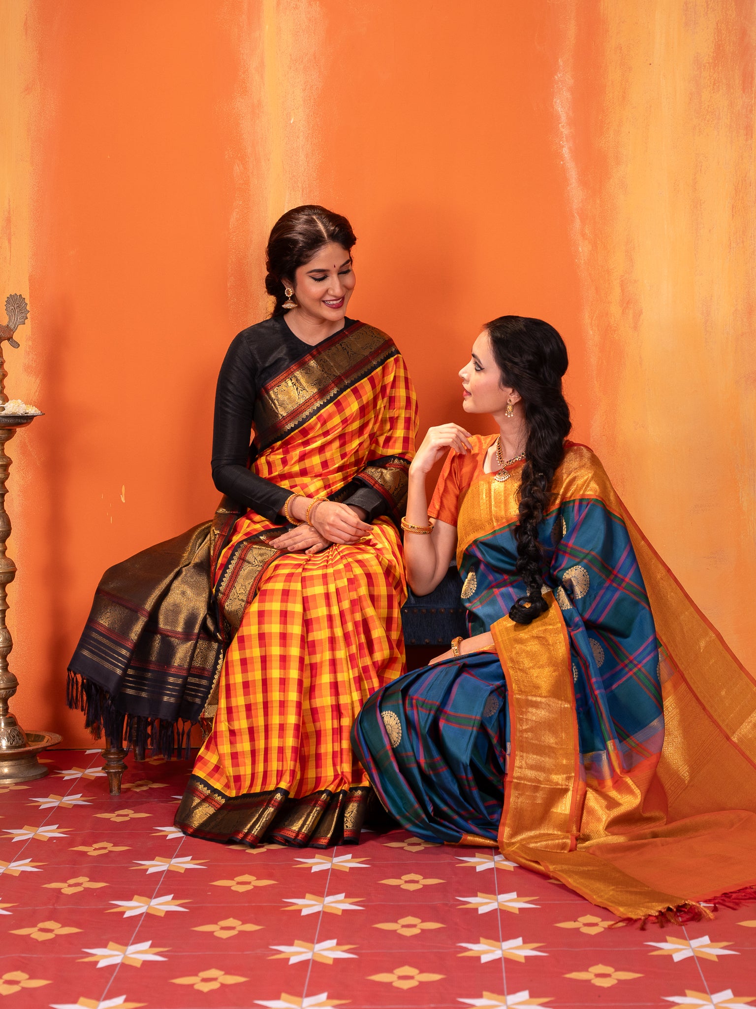 Two women in vintage traditional Kanchipuram silk sarees sitting on a patterned floor against an orange wall.