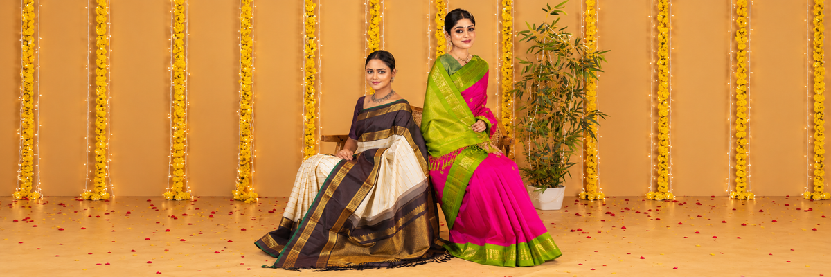 Two women seated side by side in traditional Indian Kanchipuram silk sarees against a festive backdrop of hanging yellow flowers and warm string lights. The woman on the left wears a dark brown and beige saree with gold zari borders, and the woman on the right wears a bright pink and lime green saree with a wide gold border.
