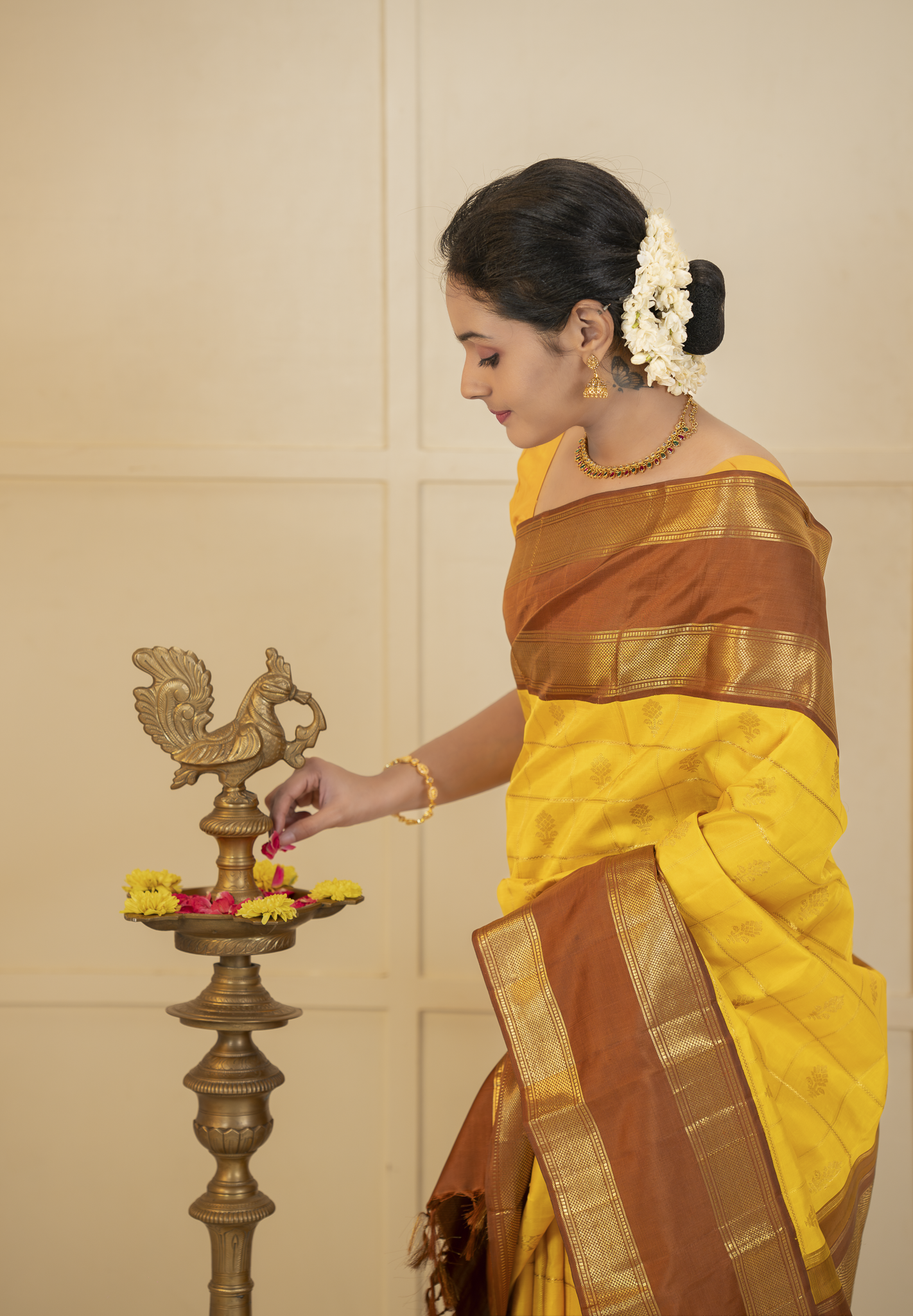 Woman in a yellow and brown saree performing a traditional ritual with a brass lamp.