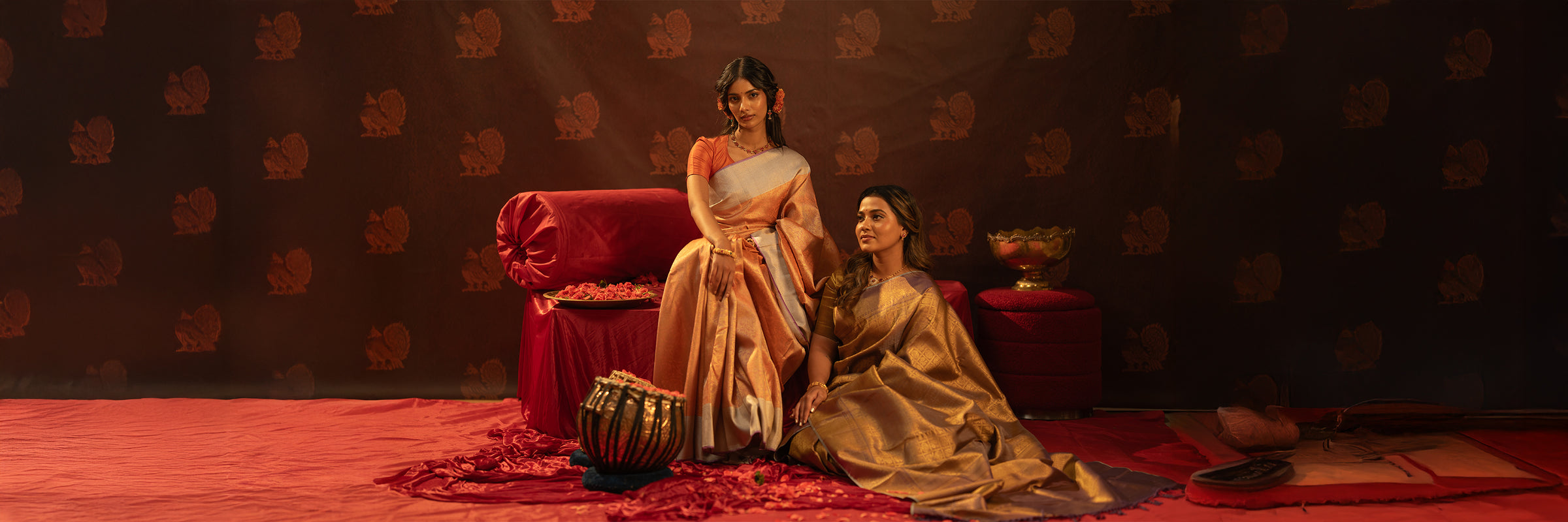Two women in traditional Kanchipuram silks sitting on a red couch against a patterned motif wall.