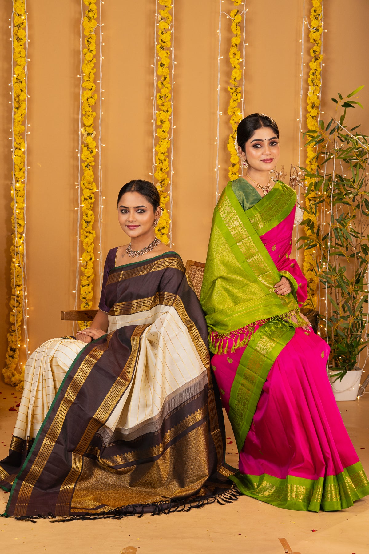 Two women seated side by side in traditional Indian Kanchipuram silk sarees against a festive backdrop of hanging yellow flowers and warm string lights. The woman on the left wears a dark brown and beige saree with gold zari borders, and the woman on the right wears a bright pink and lime green saree with a wide gold border.